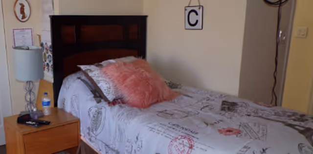 A single bed with a dark wooden headboard, covered with a white bedspread featuring various printed designs and text. There is a pink fluffy pillow and a brown pillow on the bed. Next to the bed is a wooden nightstand with a lamp, a water bottle, and some small items. The wall behind the bed is light-colored with a letter 'C' sign hanging on it.