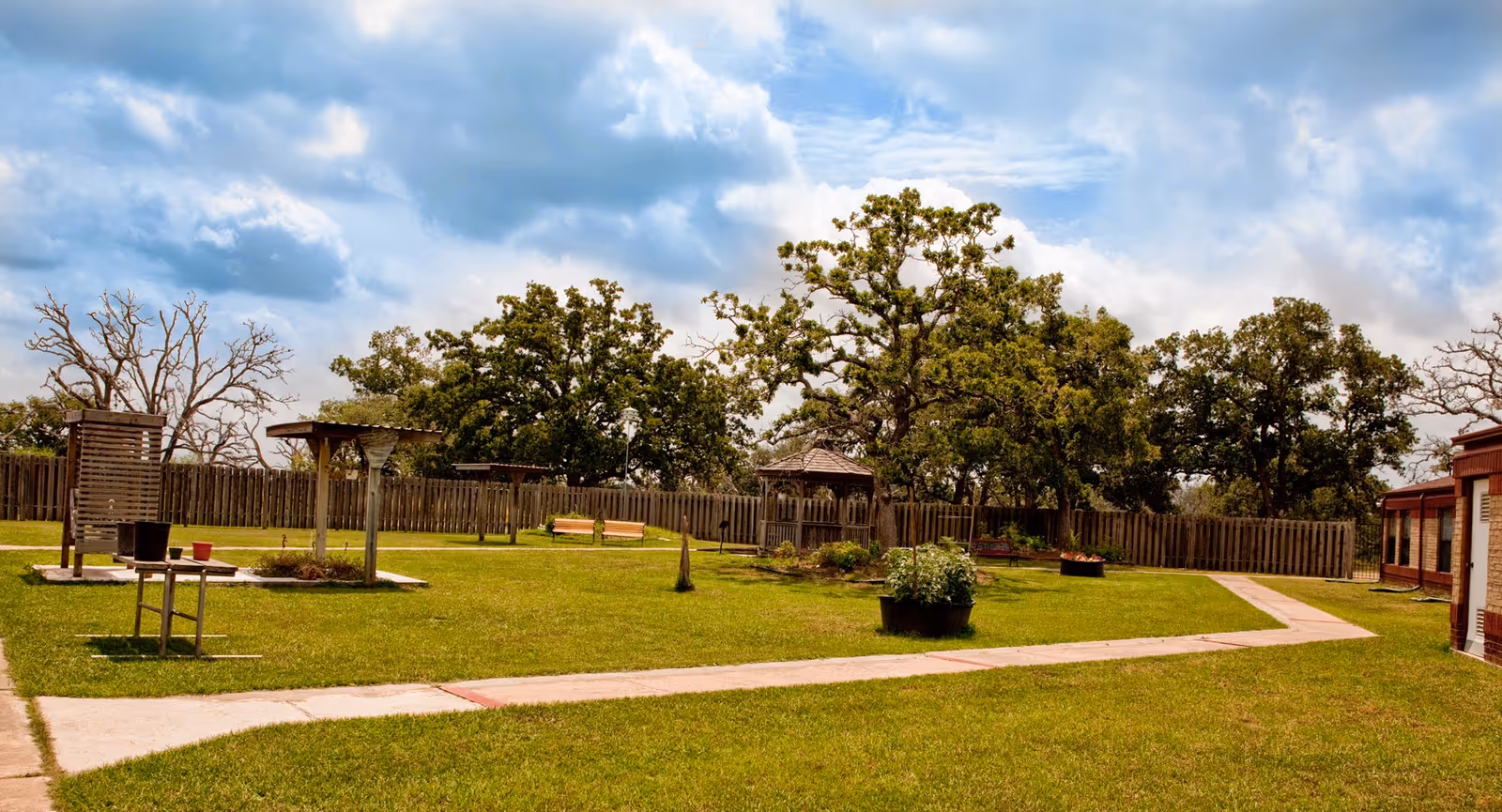 Outdoor garden area at Yoakum Nursing and Rehabilitation Center with green grass, paved walkways, wooden benches, a gazebo, and trees under a partly cloudy sky.