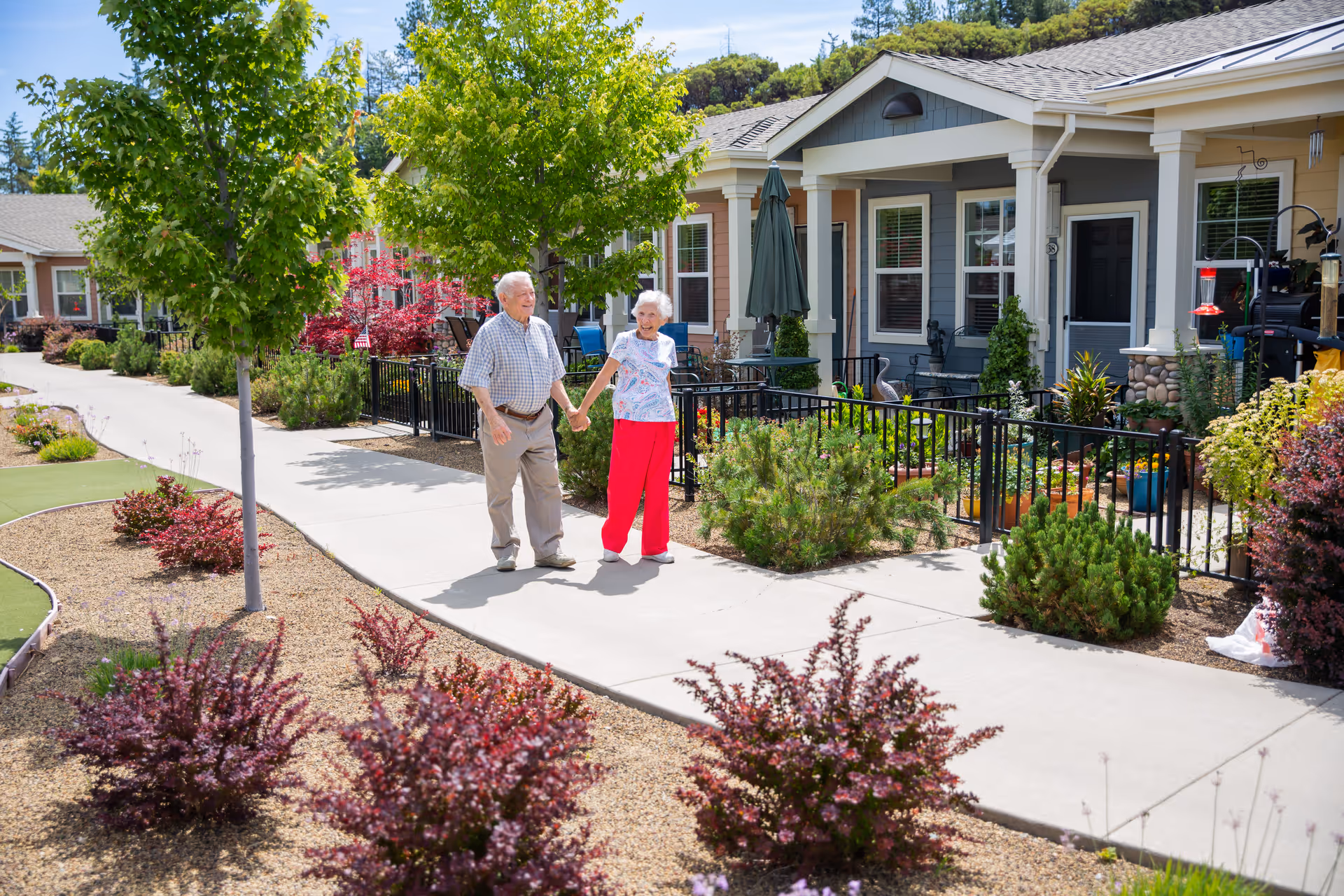 An elderly couple holding hands and walking on a paved pathway in front of single-story residential units with small fenced gardens and colorful plants at Eskaton Village Placerville.