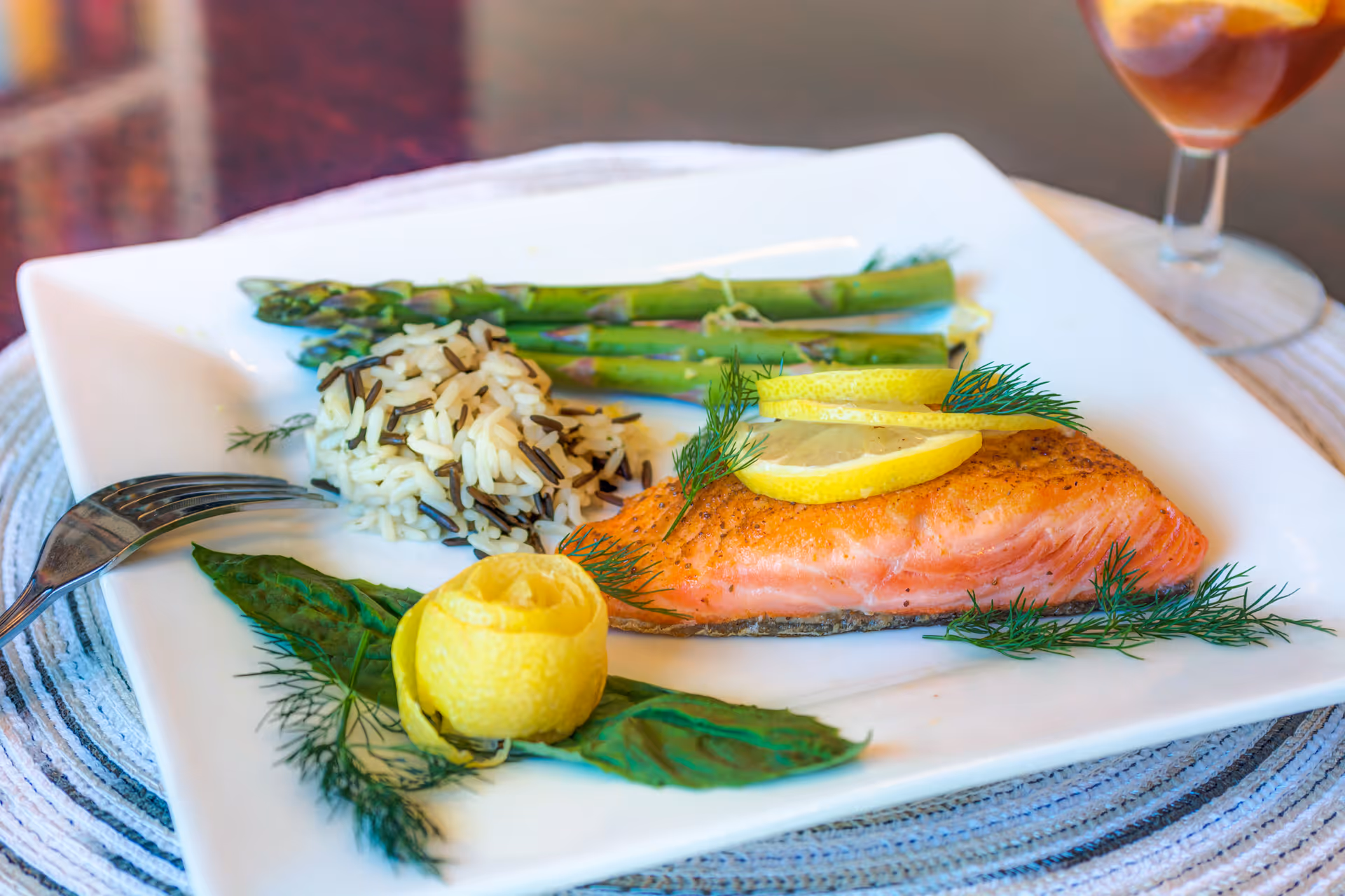 A plated meal featuring a cooked salmon fillet garnished with lemon slices and dill, served with a side of wild rice and asparagus spears on a white square plate. A fork is placed on the left side of the plate, and a glass of iced tea is visible in the background.