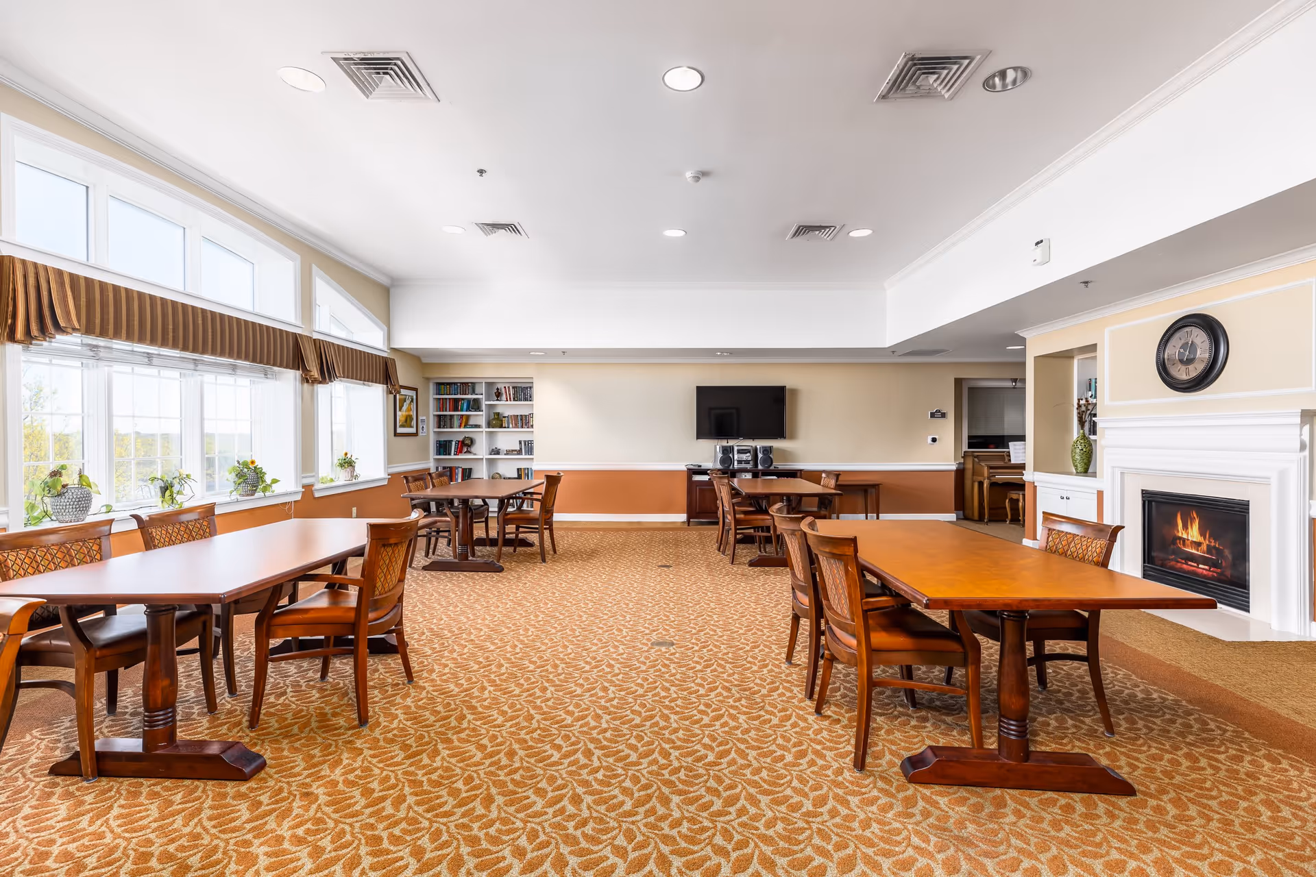 A spacious, well-lit common area with multiple wooden tables and chairs arranged on a patterned carpet. Large windows with valances let in natural light, and there are small plants on the windowsills. A fireplace with a clock above it is on the right wall, and a flat-screen TV with a stereo system is mounted on the far wall above a wooden cabinet. Bookshelves with books and framed artwork are also visible.