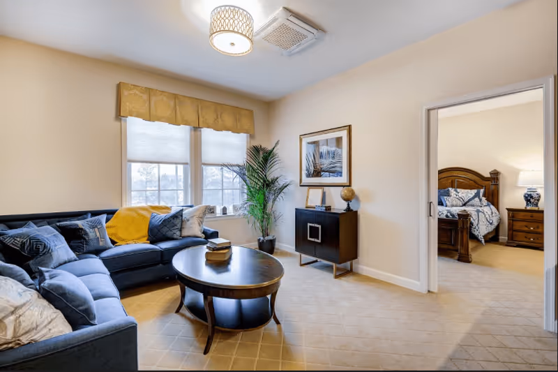 Bright furnished living room with a blue sectional sofa, round coffee table, potted plant, and a doorway opening to a bedroom.