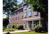 Exterior view of a multi-story brick building with large windows and a covered entrance. The building is surrounded by green trees and bushes, with a well-maintained lawn and a sign near the sidewalk.