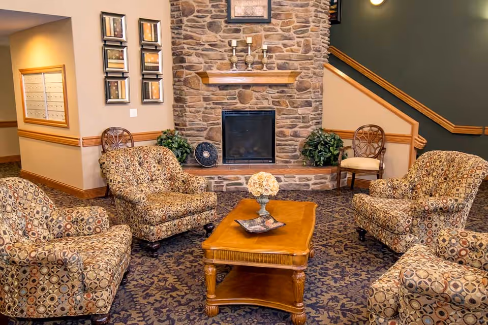 A cozy seating area in a senior living facility with four patterned armchairs arranged around a wooden coffee table. The background features a stone fireplace with decorative items on the mantel, framed artwork on the wall, and a staircase with wooden handrails. The carpet has a dark floral pattern, and there are two potted plants on either side of the fireplace.