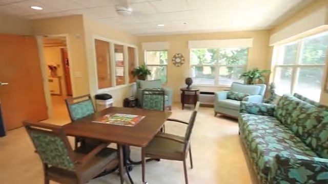 A bright living room area with large windows letting in natural light. The room features a wooden table with four chairs, a patterned green sofa, two armchairs, a small round side table with a decorative item, and plants near the windows. The walls are light-colored, and there is an open doorway leading to another room.