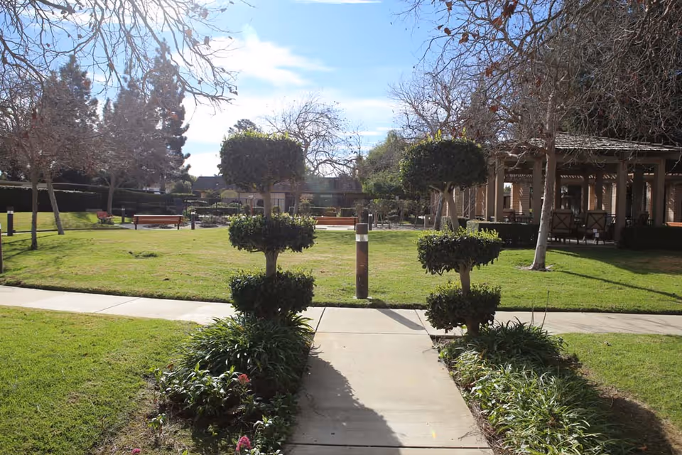 Well-kept outdoor courtyard with trimmed topiary trees flanking a central walkway, benches on the lawn, and a pergola-covered seating area under a blue sky.