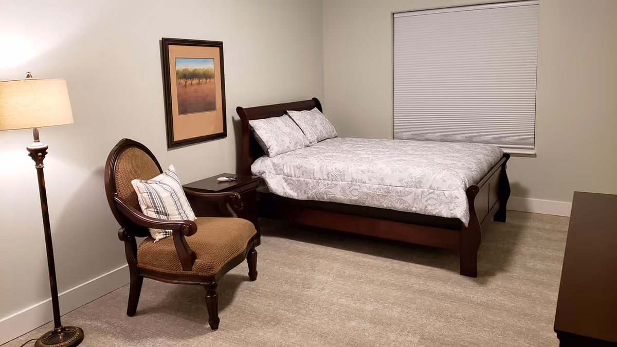 A bedroom with a wooden bed frame and white patterned bedding, a wooden nightstand with a remote control, a cushioned wooden armchair with a striped pillow, a floor lamp, a framed picture on the wall, and a window with a closed blind.
