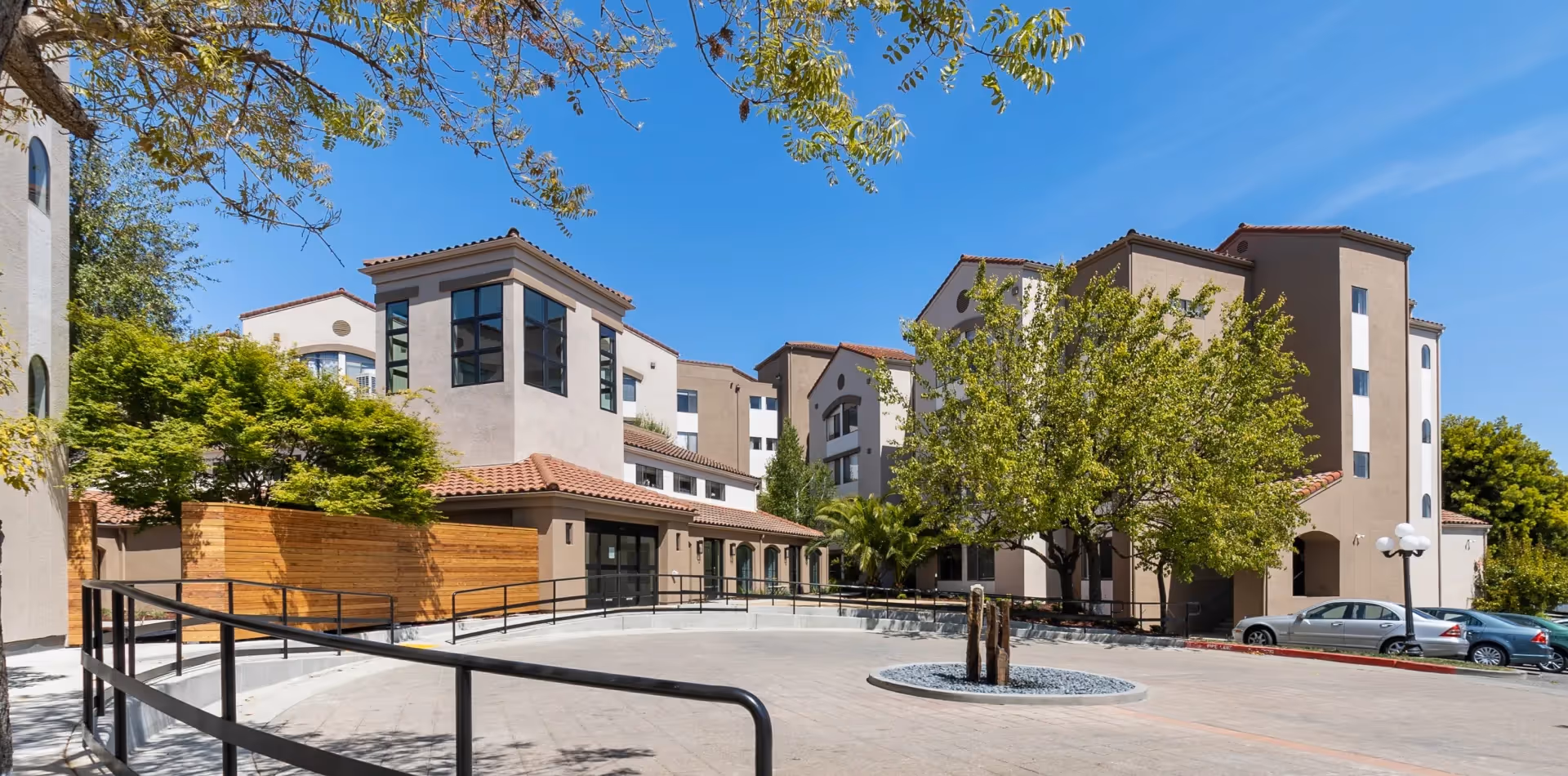 Exterior view of Cadence Millbrae senior living facility showing a multi-story building with beige walls and red-tiled roofs, surrounded by trees and a clear blue sky. There is a circular driveway with a small stone and wood sculpture in the center and several parked cars to the right.