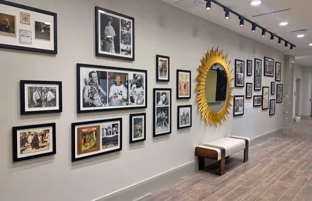 A hallway with a gallery wall featuring multiple framed photographs and a large decorative sunburst mirror. Below the mirror is a bench with a cushioned seat. The floor has wood-like tiles and the ceiling has track lighting.