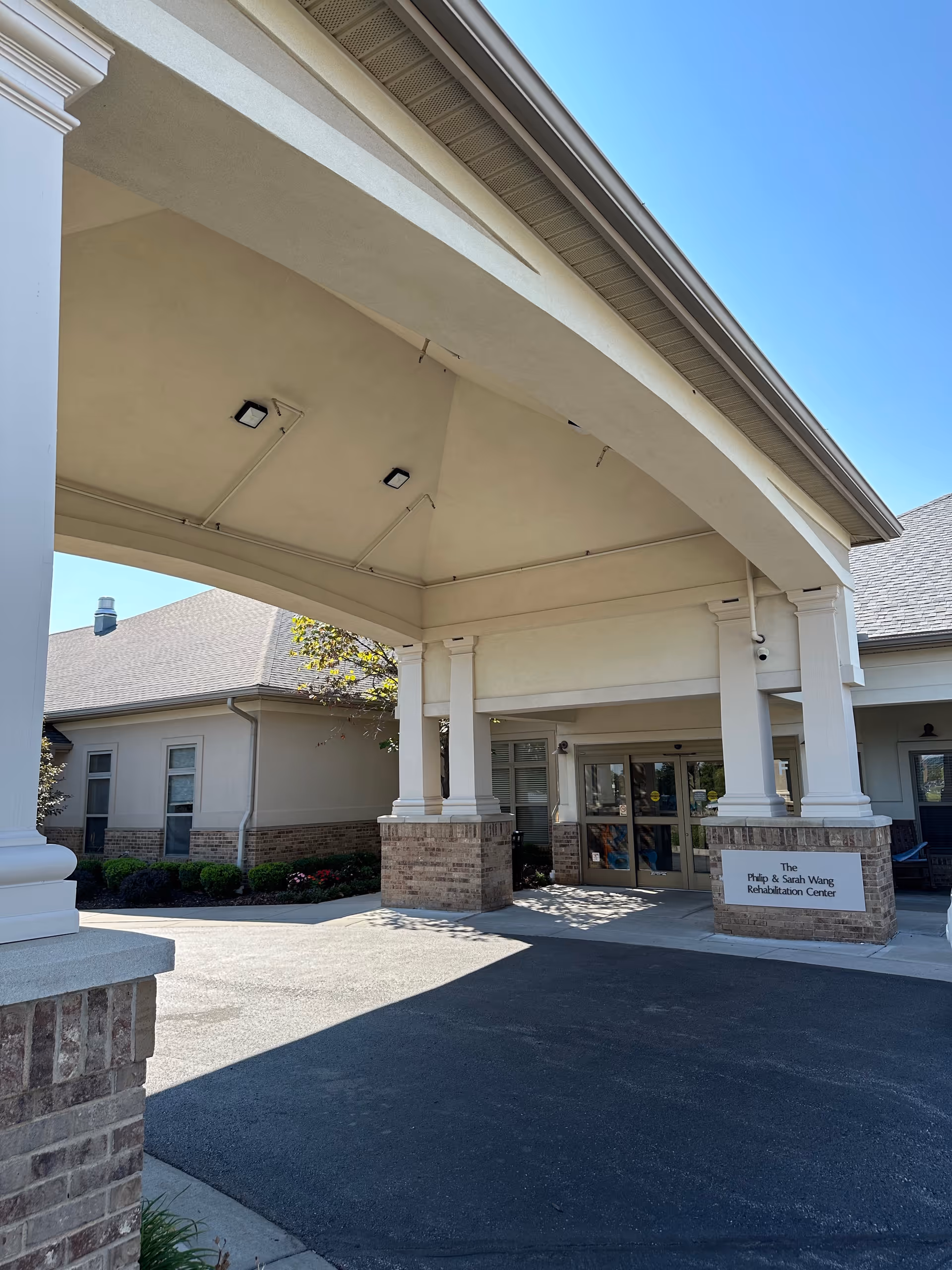 Covered entrance/portico of a senior living facility with brick pillars and automatic glass doors under a clear blue sky.