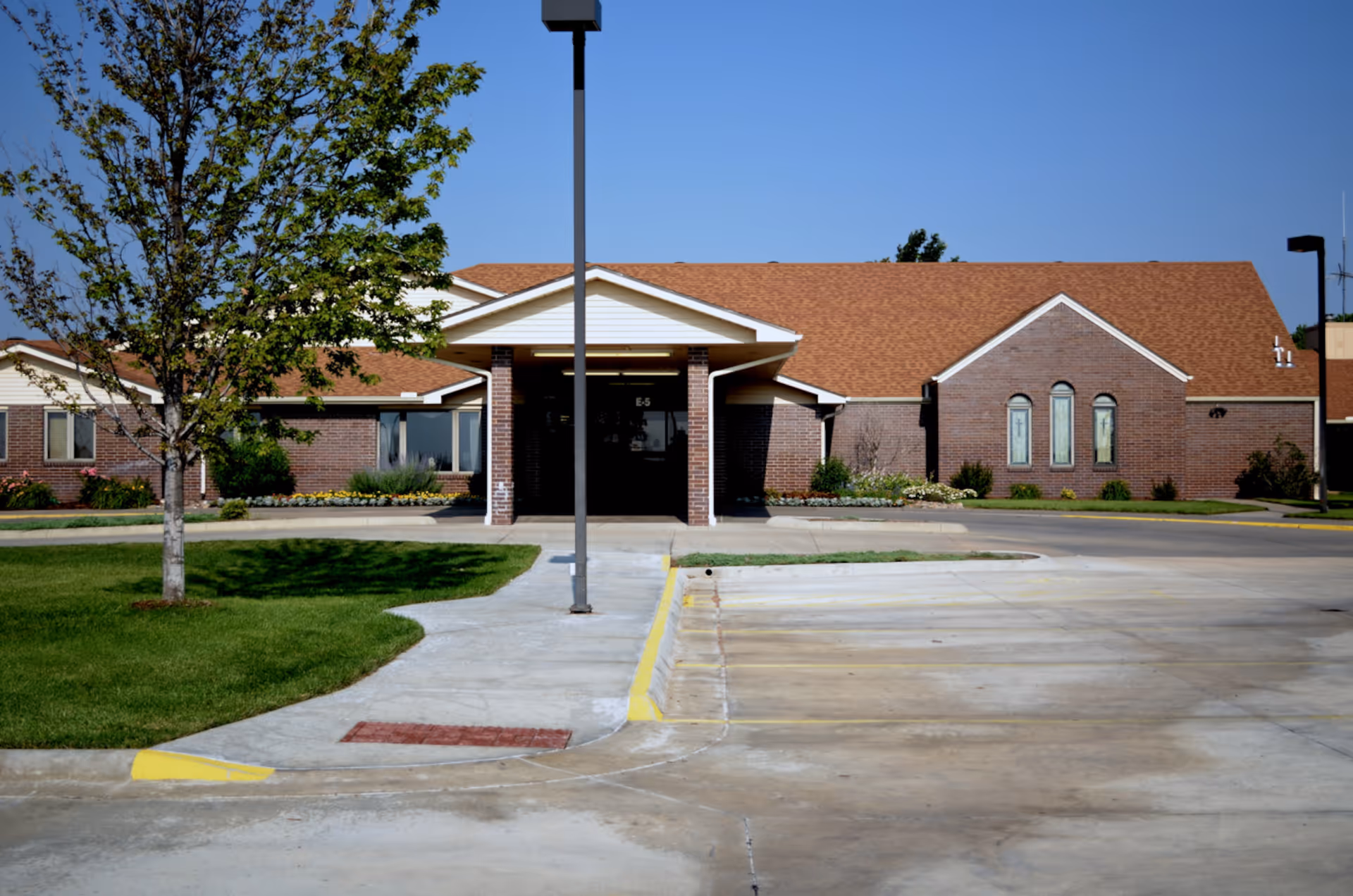 Exterior view of a single-story brick building with a brown shingled roof, a covered entrance supported by brick columns, a tree and green lawn on the left, and a concrete parking area in front under a clear blue sky.