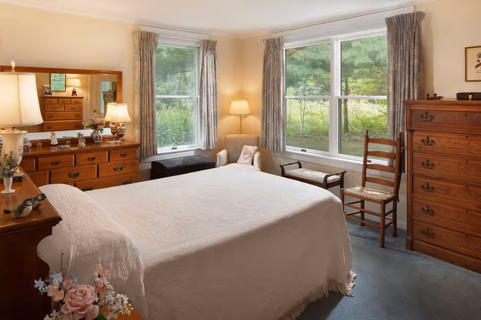 Sunlit bedroom with a neatly made bed, wooden dressers and chairs, and two large windows overlooking greenery.