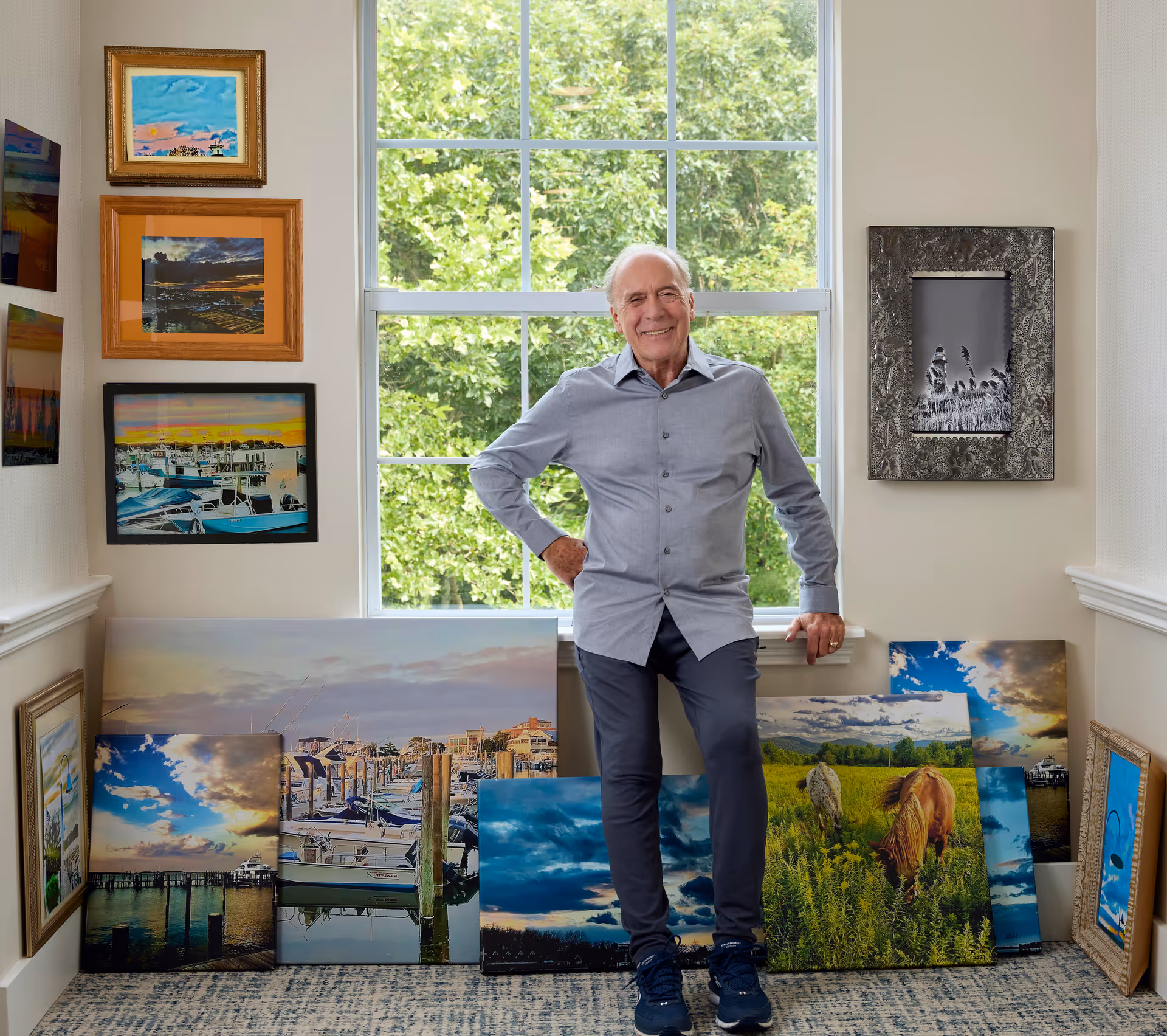 An elderly man standing and smiling in front of a large window with green trees outside. He is surrounded by various framed paintings and photographs leaning against the wall and hung on the walls, depicting nature scenes, boats, and landscapes.