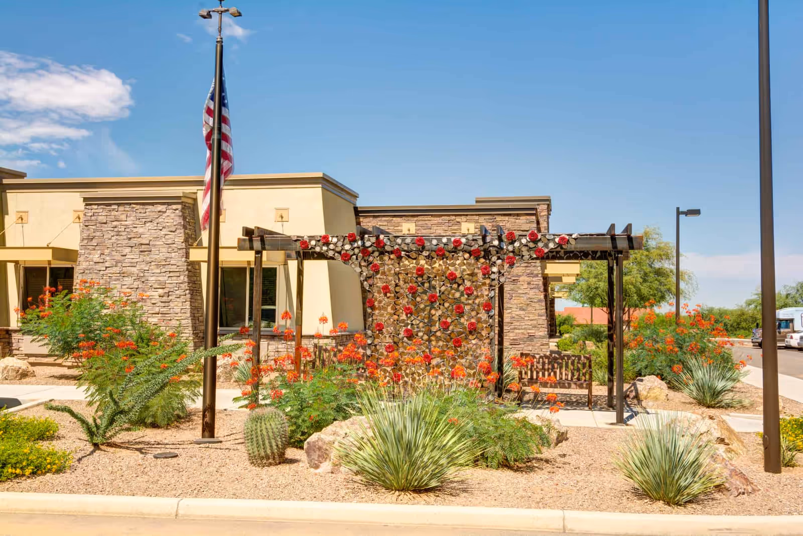 Exterior view of Catalina Springs Memory Care facility featuring a landscaped garden with desert plants, a decorative metal wall with red flower accents, a wooden bench, and an American flag on a flagpole under a clear blue sky.