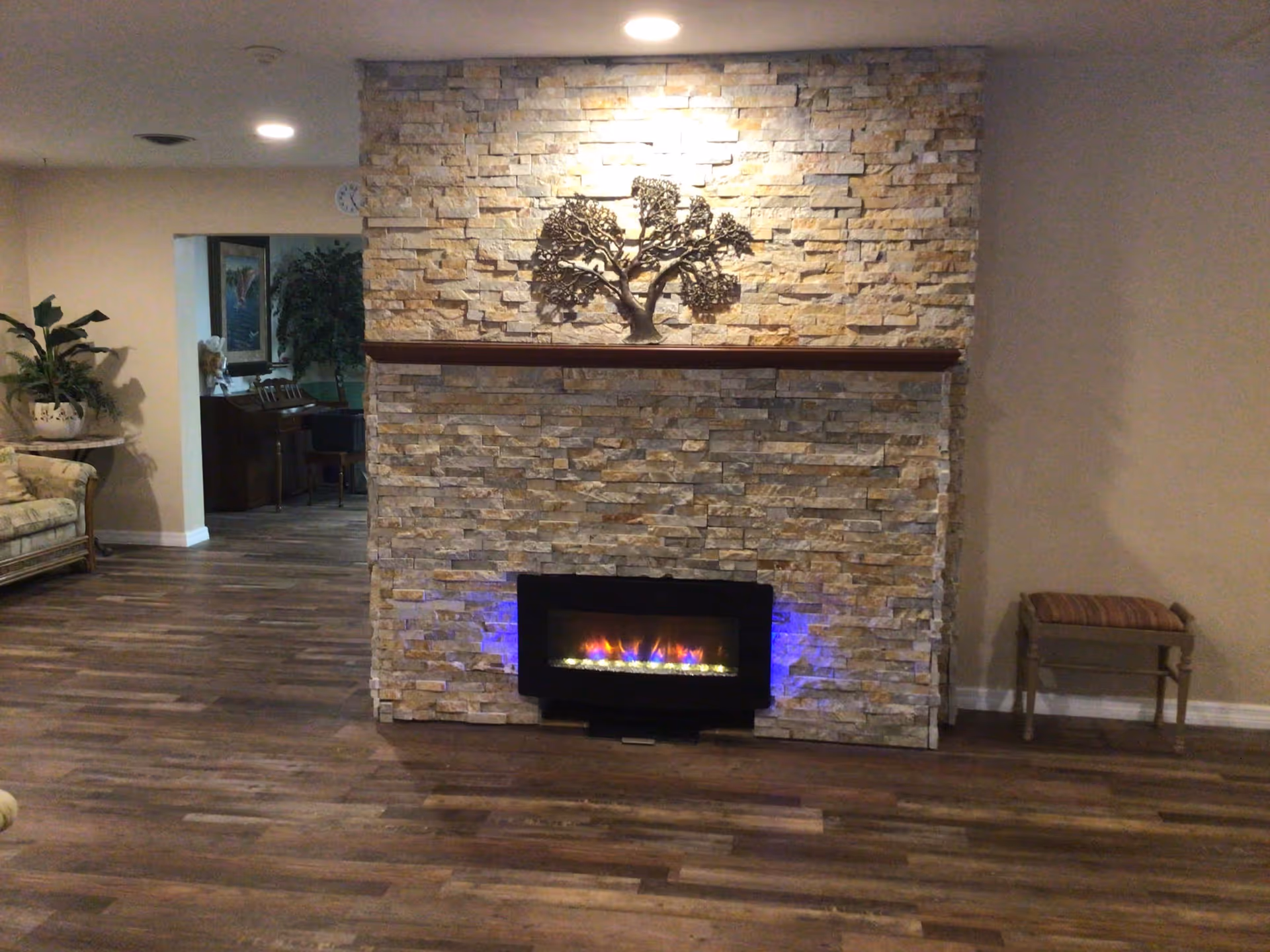 A cozy living room area with a stone fireplace featuring a modern electric insert with blue and orange flames. Above the fireplace is a wooden mantel with a decorative metal tree sculpture mounted on the stone wall. The room has wood flooring and neutral-colored walls. To the left, there is a floral-patterned sofa and a round side table with a potted plant. In the background, a piano and additional plants are visible in an adjacent room. On the right side, there is a small wooden bench with a cushioned seat.