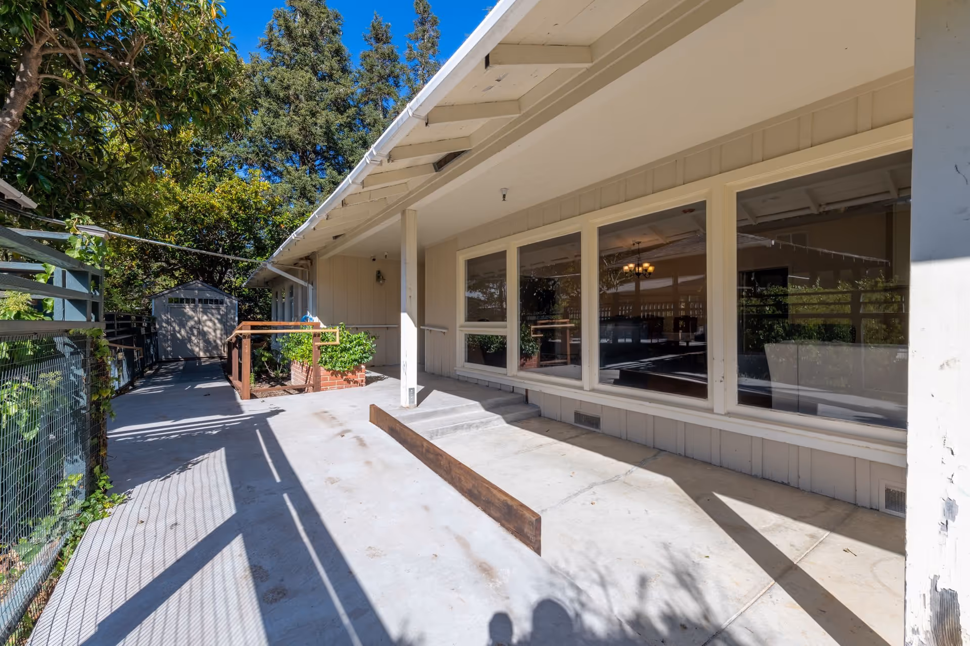 Outdoor patio area of Marin Terrace Assisted Living with a concrete walkway, a small ramp with wooden railing, a few steps leading up to a covered porch with large windows, and a garden shed in the background surrounded by trees and greenery under a clear blue sky.