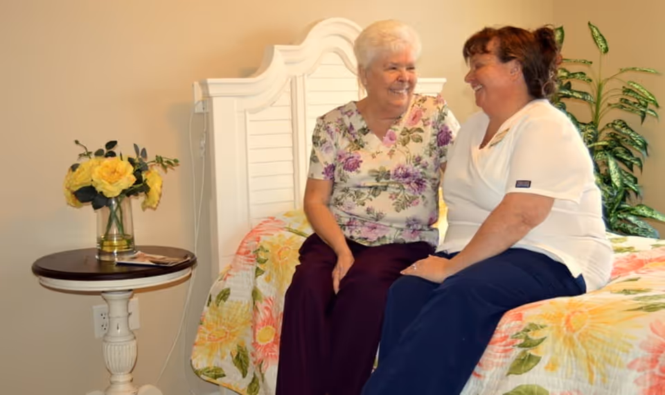 An elderly woman and a caregiver sitting on a bed in a bedroom, smiling and engaging in a friendly conversation. The bed has a floral quilt, and there is a small round table with a vase of yellow flowers next to the bed. A green leafy plant is visible in the background.