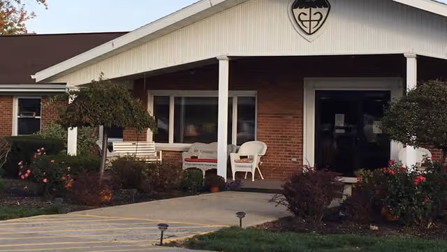 Front exterior view of a single-story brick building with a covered entrance supported by white columns. There are white chairs and a bench on the porch, surrounded by landscaped bushes and flowers. A shield emblem is visible near the roof peak above the entrance.
