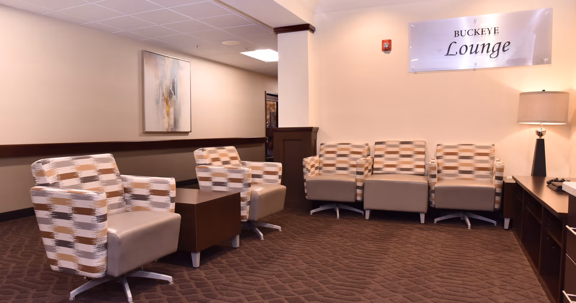 A lounge area with five patterned armchairs arranged around two small wooden tables. The walls are beige with a dark brown trim, and there is a framed abstract painting on one wall. A sign on the wall reads 'BUCKEYE Lounge'. A table lamp is placed on a wooden cabinet to the right.