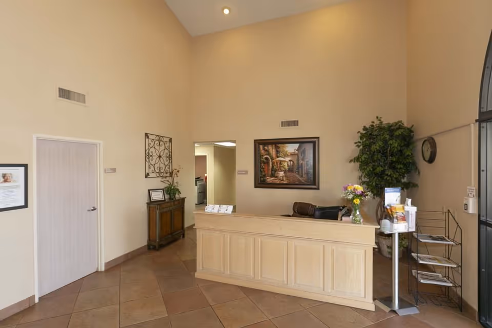 Reception area of Mountain View Care Center with a wooden front desk, a vase of flowers, a framed painting on the wall, a potted plant, a clock, and a brochure stand. The walls are beige and the floor is tiled.