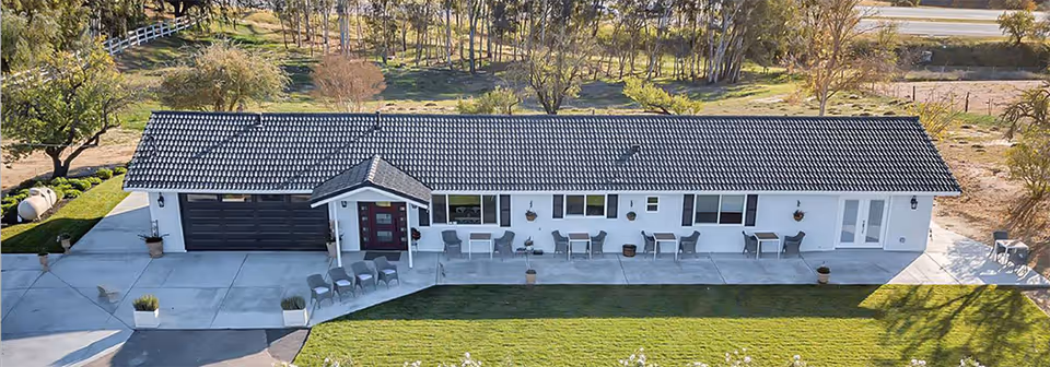 Aerial view of a single-story building with a dark tiled roof and white exterior walls. The building has a covered entrance with a red door, multiple windows, and several outdoor seating areas with tables and chairs on a concrete patio. The surrounding area includes green grass, trees, and a paved driveway.