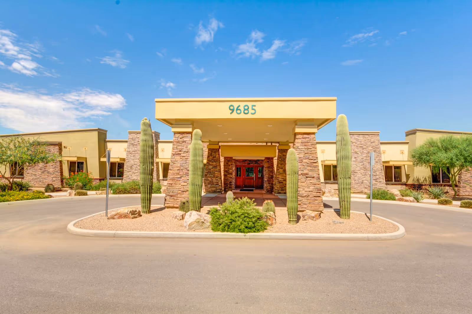 Front exterior view of Catalina Springs Memory Care facility with a covered entrance supported by stone pillars, surrounded by desert landscaping including tall cacti and shrubs under a clear blue sky.