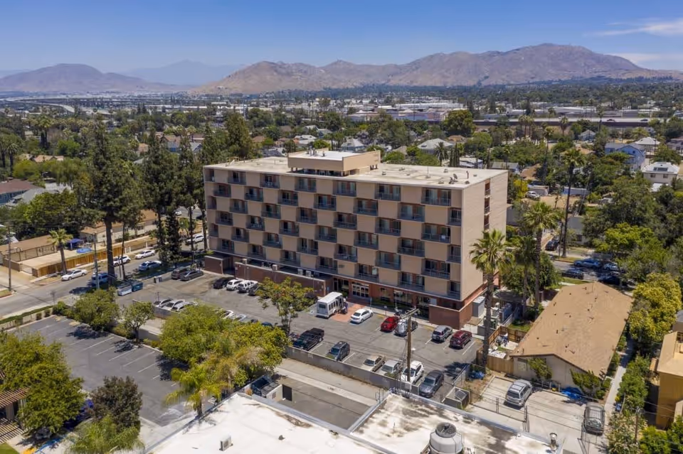 Aerial view of a six-story senior living building with balconies and a parking lot, set in a residential neighborhood with trees and distant mountains.