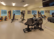 A fitness room with exercise equipment including stationary bikes, weight machines, and dumbbells. The room has large windows, mirrors on one wall, and bright overhead lighting.