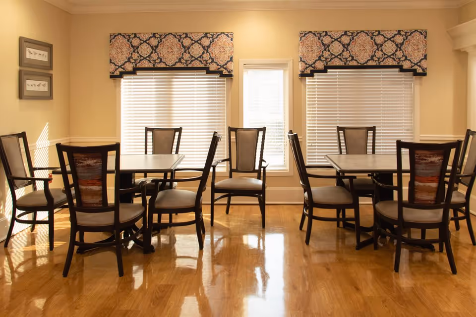 A dining area with two square tables, each surrounded by four wooden chairs with cushioned seats and backs. The room has polished wooden floors, two large windows with white blinds and decorative valances, and a smaller window in between. The walls are painted a light beige color and there are two framed pictures on the left wall.