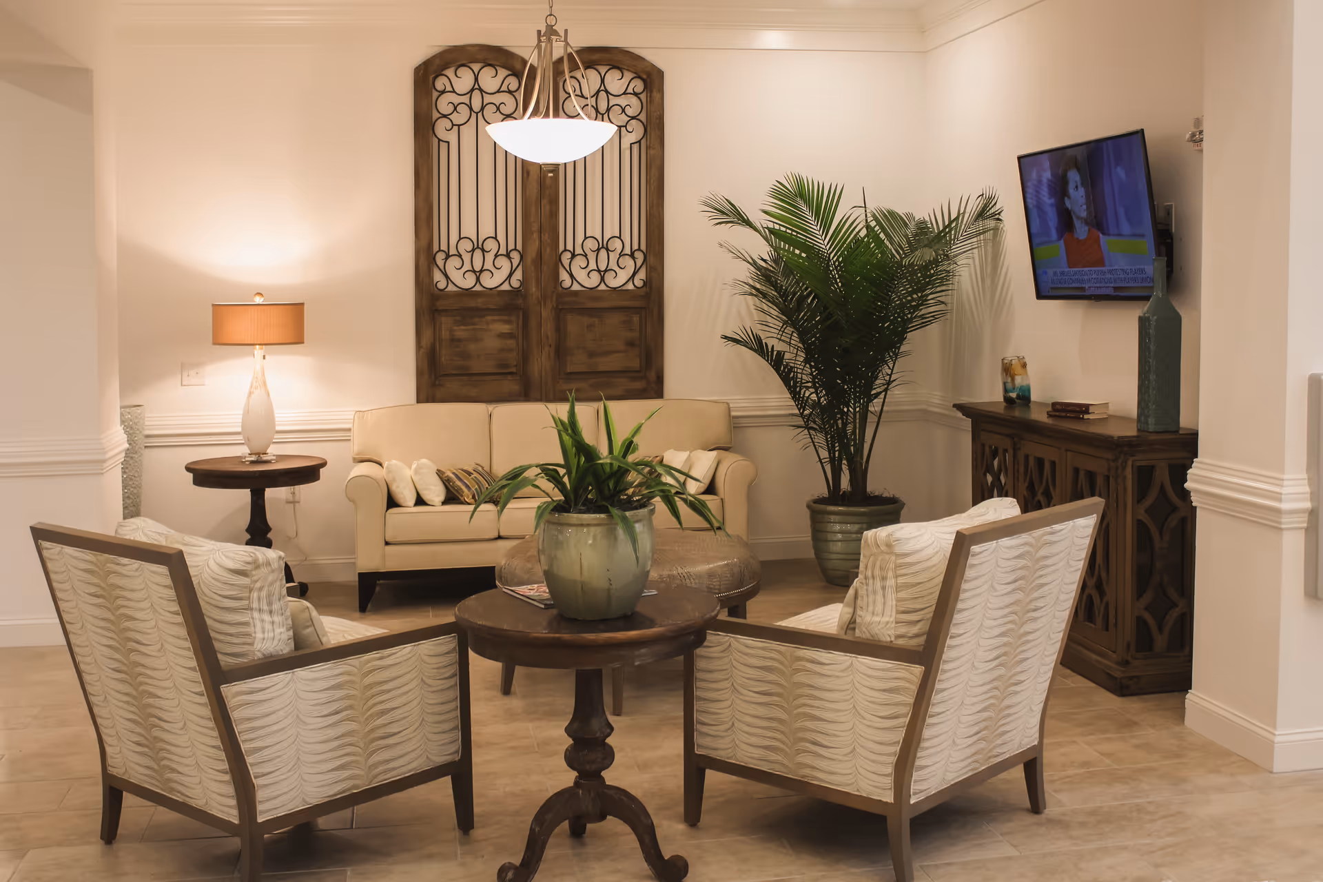 A cozy living room area with a beige sofa, two patterned armchairs, a round wooden coffee table with a potted plant, a side table with a lamp, a large potted plant in the corner, a wall-mounted TV, and decorative wooden wall art behind the sofa.
