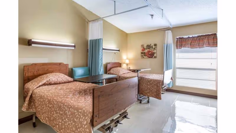 A nursing facility room with two hospital-style beds covered in brown patterned bedspreads. Each bed has a small overbed table and a privacy curtain hanging from the ceiling. The room has beige walls, a large window with blinds and a valance, a small lamp on a side table, and a floral wall decoration.