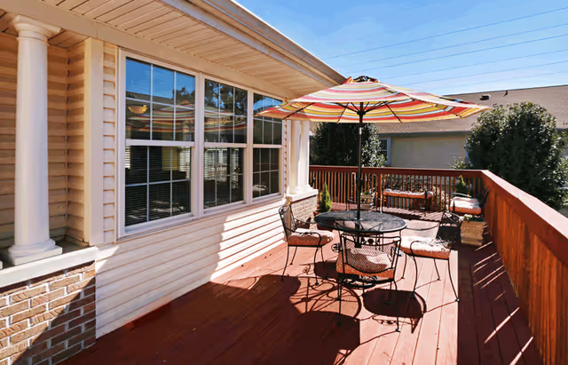 Outdoor wooden deck area with a round metal table and four chairs, each with cushions. A large striped umbrella provides shade over the table. The deck is attached to a beige building with large windows and white columns. There is a wooden railing around the deck and some greenery in the background under a clear blue sky.