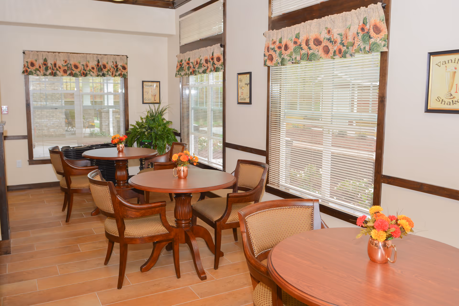 A bright dining area with round wooden tables and cushioned wooden chairs. Each table has a small copper vase with colorful flowers. The room has large windows with blinds and sunflower-patterned valances. There are framed vintage-style posters on the walls and a green potted plant in the corner.