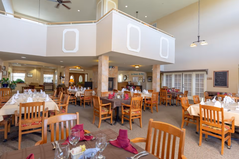 A spacious dining room in a senior living facility with multiple wooden tables and chairs arranged neatly. Tables are set with white and burgundy napkins, glassware, and cutlery. The room features high ceilings, a ceiling fan, and a fireplace in the center supported by tiled columns. Large windows and glass doors allow natural light to fill the space, and there is a chalkboard menu on one wall.