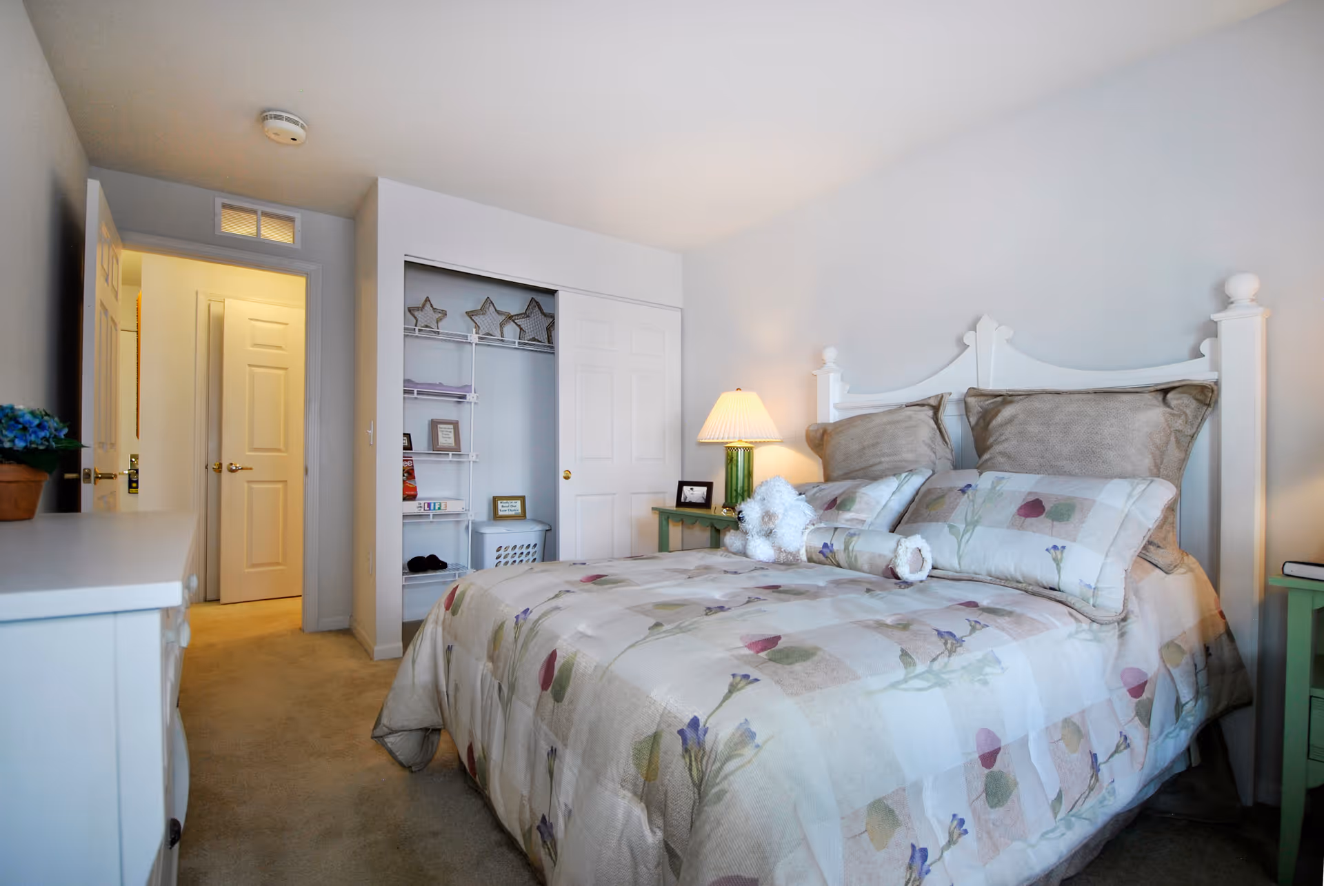A cozy bedroom with a bed featuring a floral patterned comforter and pillows, a white headboard, a green bedside table with a lamp, an open closet with shelves and decorative items, a white dresser with a potted plant, and a door leading to a hallway.