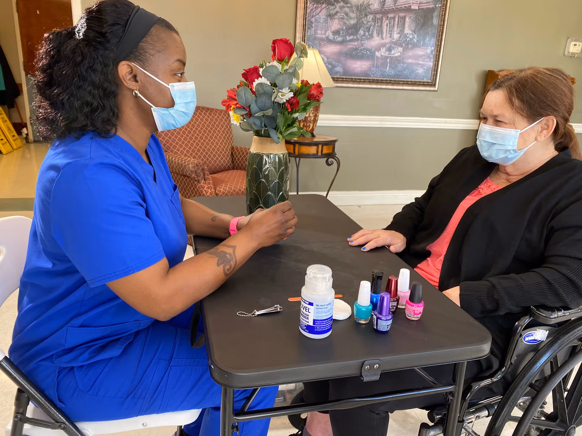 A healthcare worker in blue scrubs and a face mask is painting the nails of a woman in a wheelchair, who is also wearing a face mask. They are seated at a black table with several bottles of nail polish and a bottle of lotion. Behind them is a vase with red and white flowers, a lamp, a framed painting, and a patterned armchair.