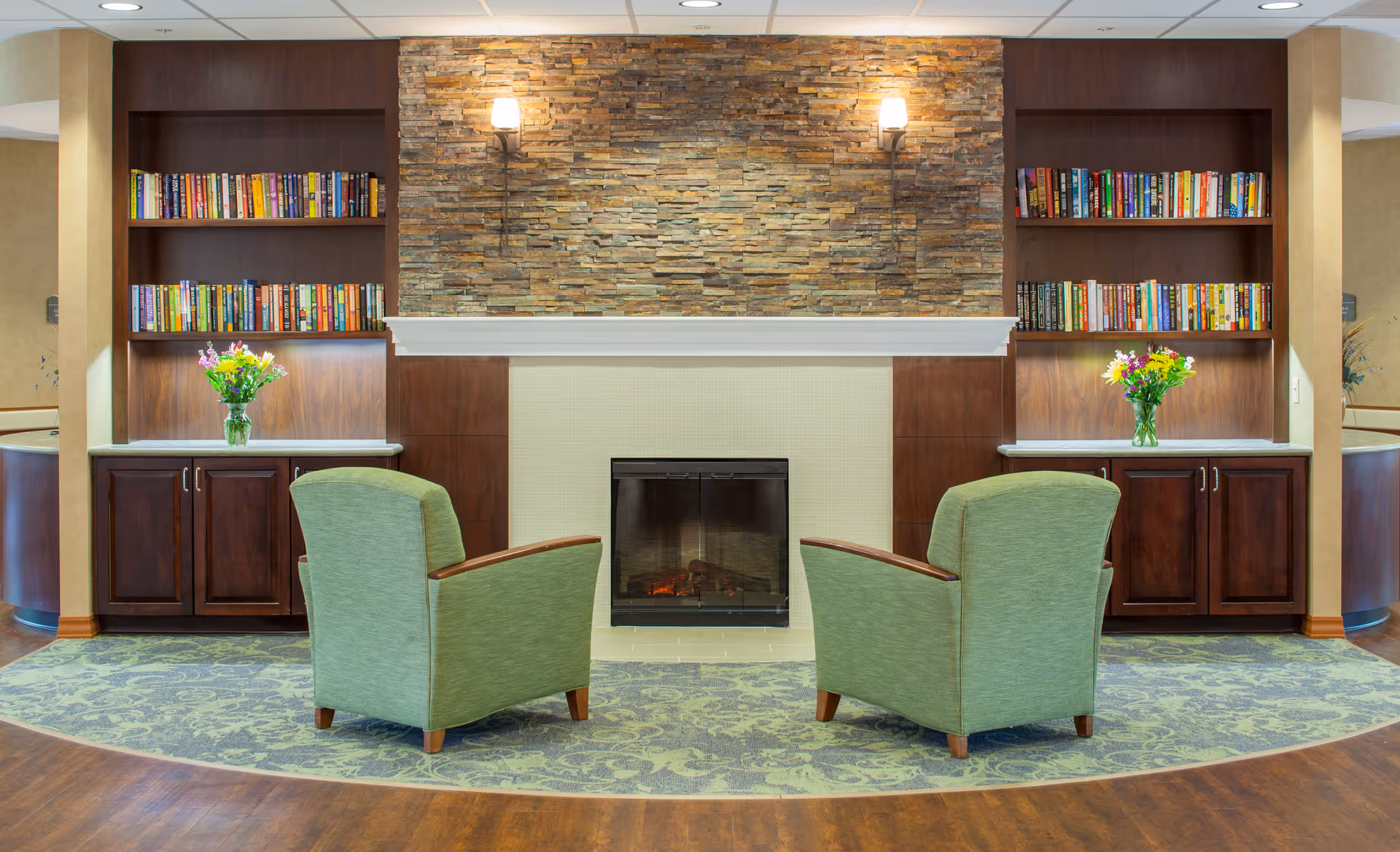 A cozy sitting area with two green armchairs facing a fireplace with a stone accent wall. On either side of the fireplace are built-in wooden bookshelves filled with books and cabinets below. Each cabinet has a vase with colorful flowers on top. The floor has a patterned carpet in front of the fireplace and wood flooring around it.