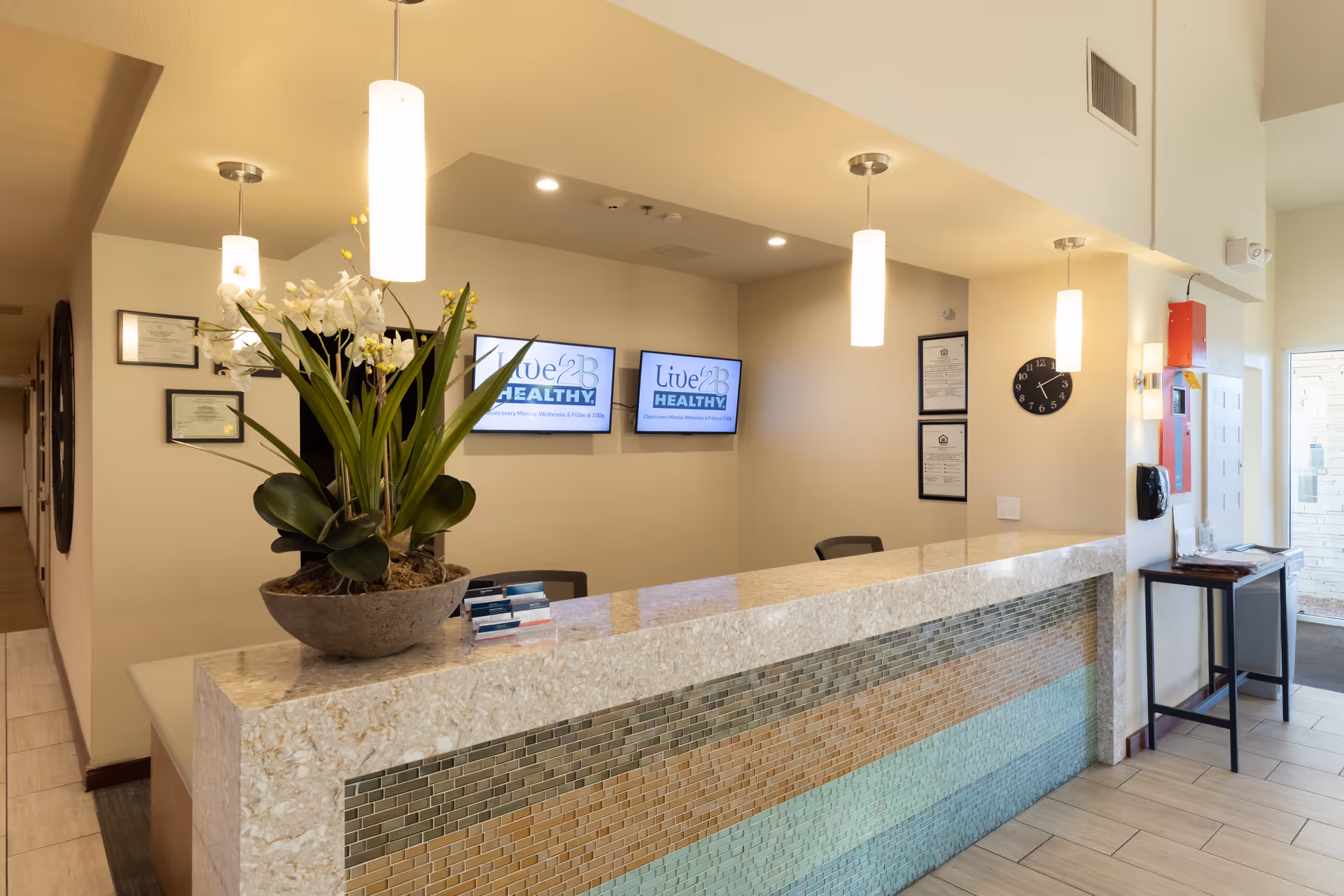 Reception area with a marble and mosaic tiled front desk, a large flower arrangement on the counter, three hanging pendant lights, two wall-mounted screens displaying 'Live 2B Healthy', a clock on the wall, framed certificates, and a hallway leading to other rooms.