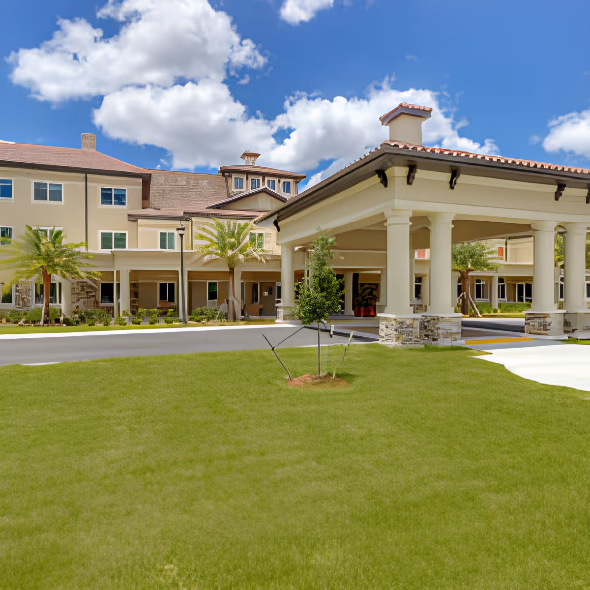 Front entrance and covered porte-cochere of a multi-story senior living building with a manicured lawn and palm trees under a blue sky.
