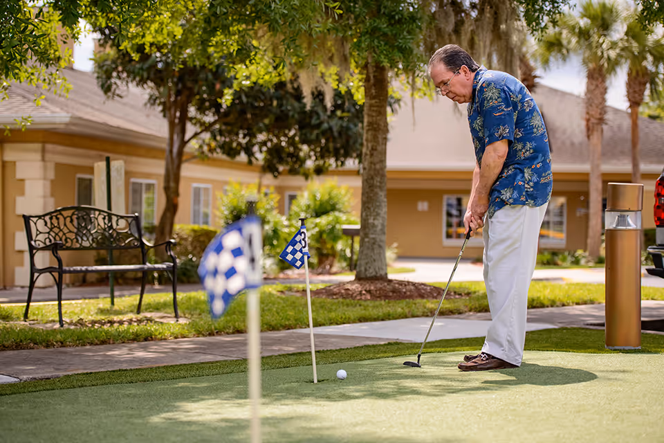 An elderly man wearing a blue floral shirt and white pants is playing mini golf on a putting green outside. There are small checkered flags on the green, a bench, trees, and a building in the background under a sunny sky.