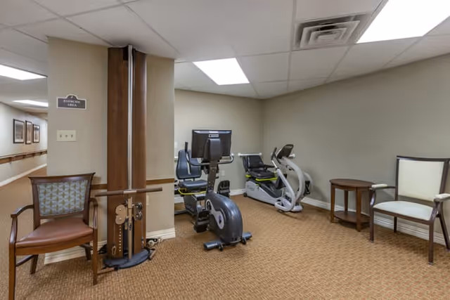 Small exercise room with stationary bikes, a multi-use fitness machine and chairs in a carpeted interior space.