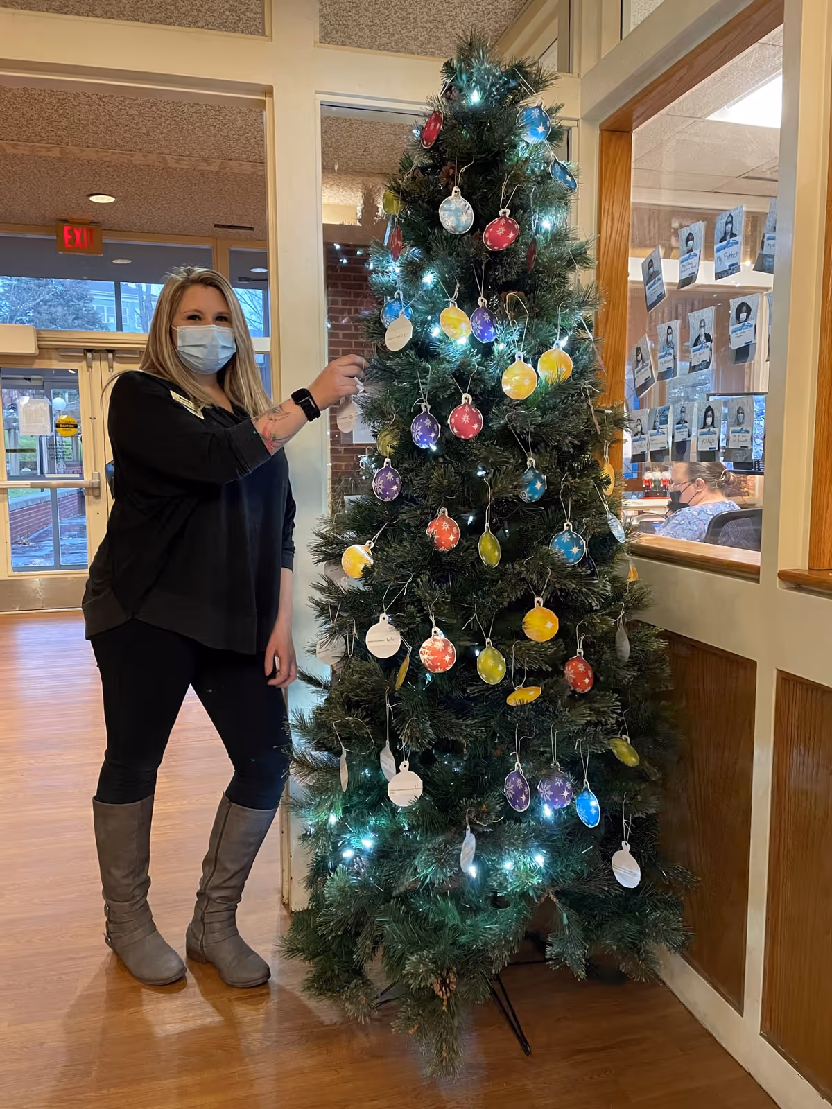 A woman wearing a face mask, black top, black pants, and gray boots stands next to a decorated Christmas tree indoors. The tree is adorned with colorful round ornaments and white lights. Behind her, there is a glass window with photos displayed and a person sitting at a desk. The setting appears to be a communal area in a facility.