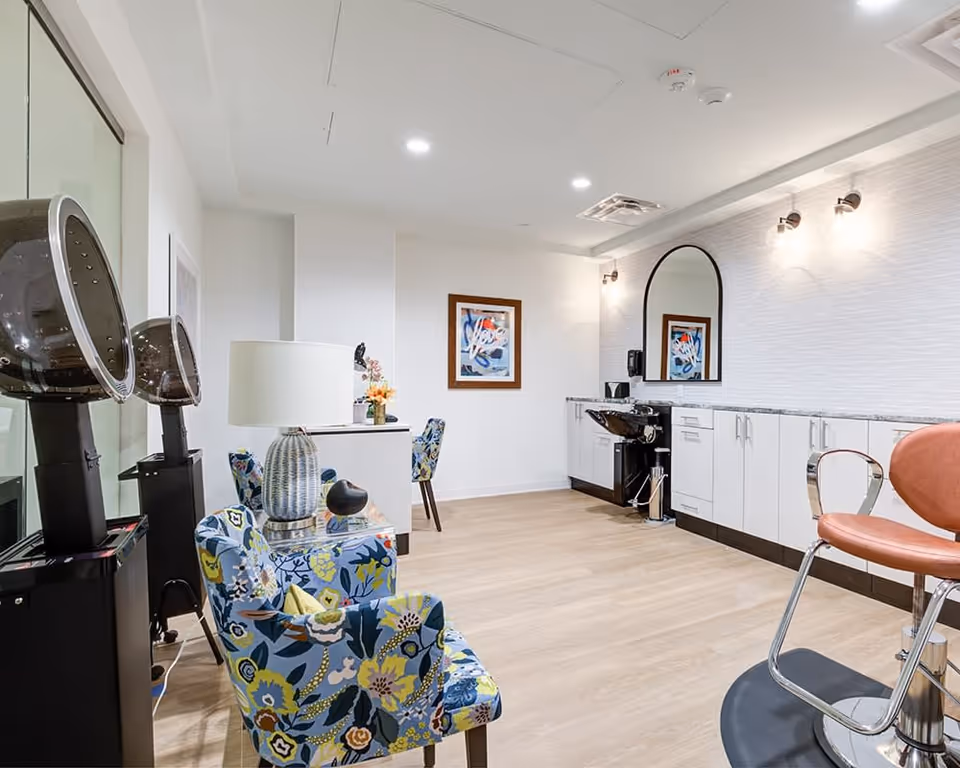 Interior of a modern salon room with floral patterned chairs, two black hair dryers, a lamp on a small table, a large mirror on the wall, white cabinets with a black sink, and a peach salon chair on a wooden floor.
