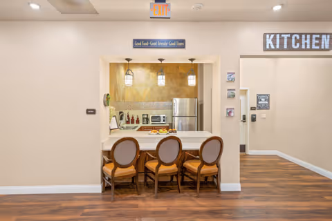 Interior view of a kitchen area in a senior living facility with three wooden chairs with cushioned seats placed at a counter. The kitchen has a stainless steel refrigerator, microwave, and various kitchen items on the counter. Above the kitchen window is a sign that reads 'Good Food - Good Friends - Good Times'. To the right, a large sign on the wall says 'KITCHEN'. The floor is wooden, and the walls are painted beige.