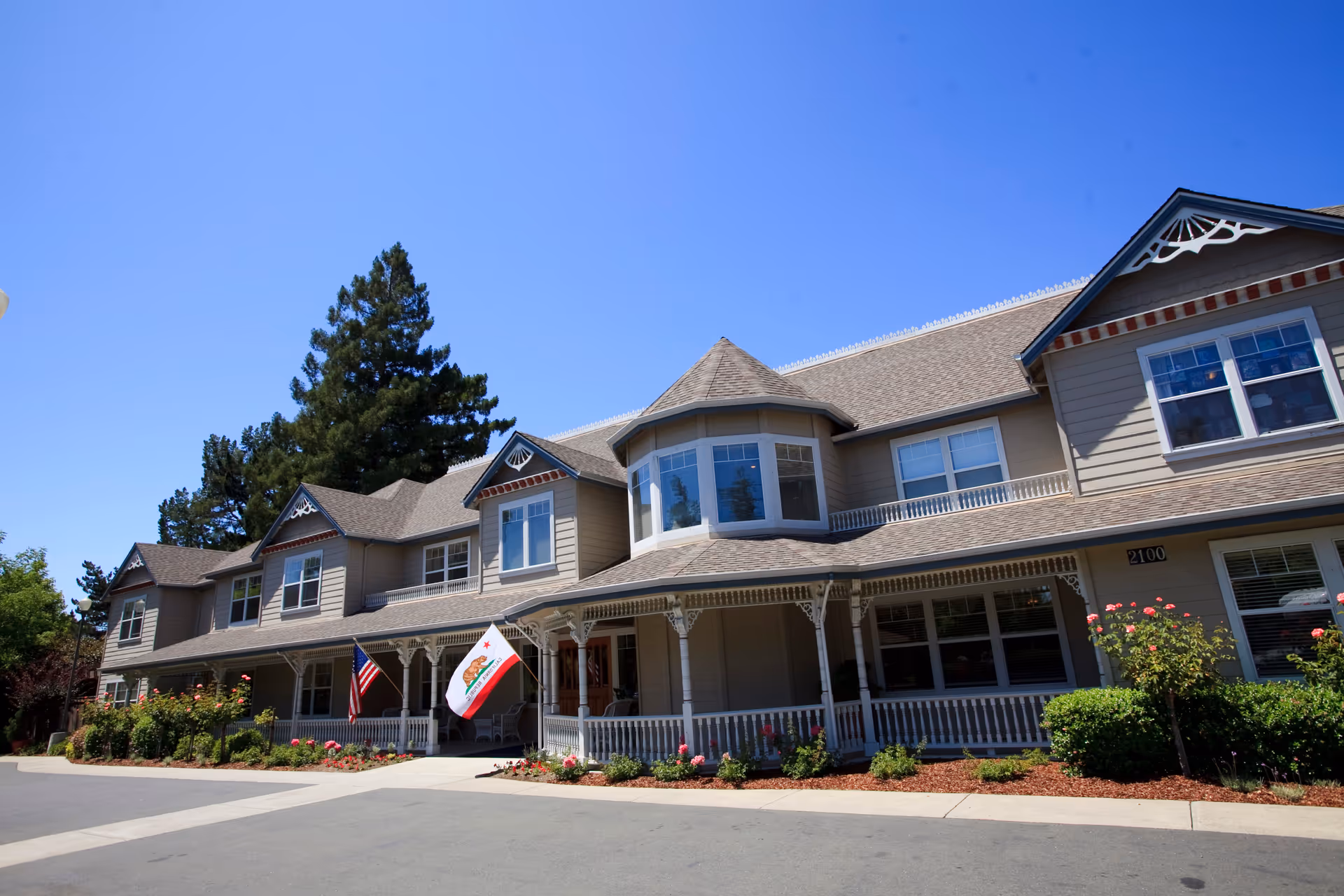 Front exterior of a two-story Victorian-style senior living building with a wraparound porch, landscaping, and American and California flags.