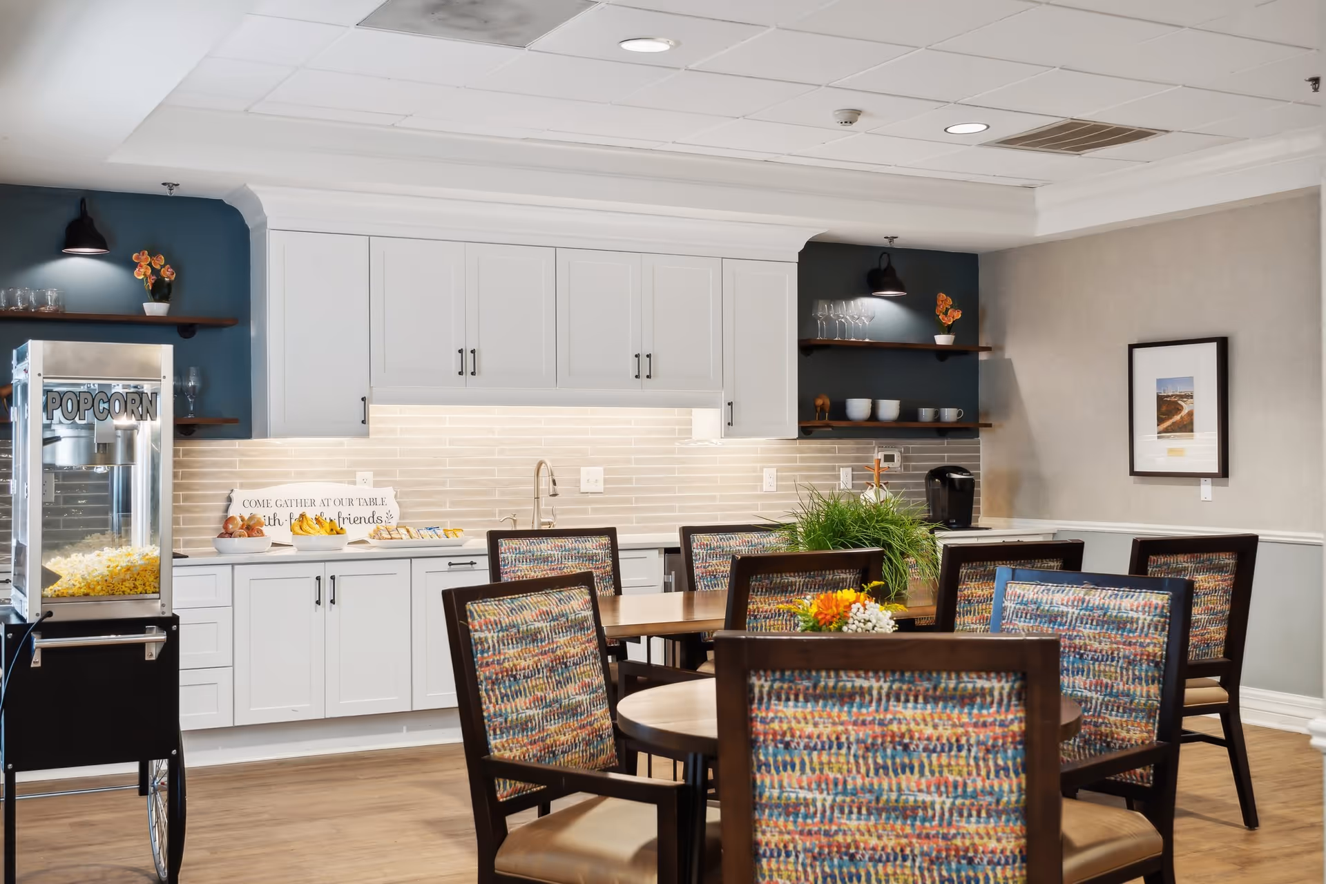 Communal dining area with round tables and patterned chairs in front of a kitchenette with white cabinets and a popcorn machine.