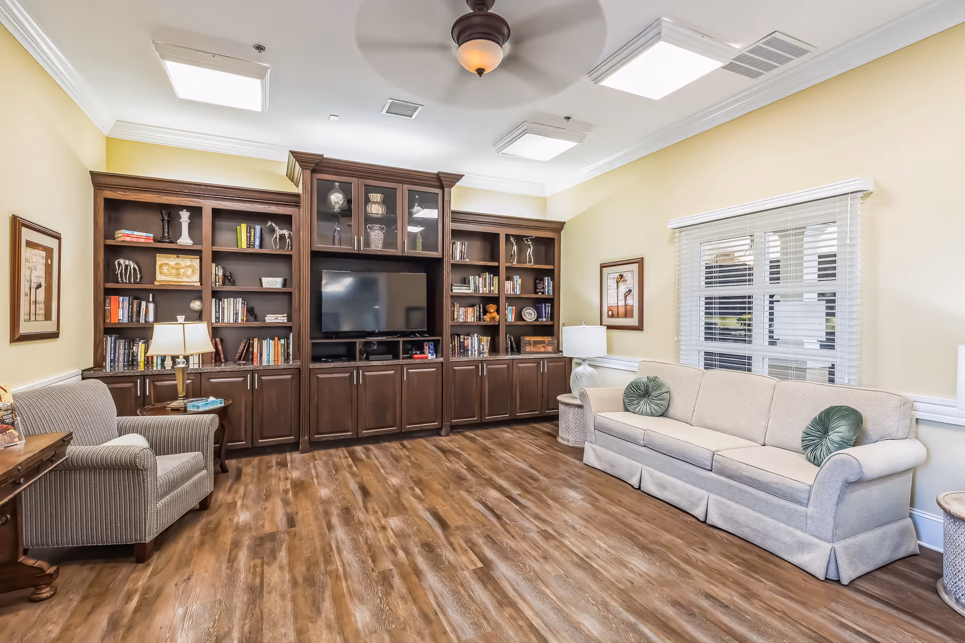 A bright and spacious living room with wooden flooring, a large built-in dark wood entertainment center with shelves filled with books and decorative items, a flat-screen TV in the center, a beige sofa with two green round pillows, a striped armchair, two side tables with lamps, framed artwork on the walls, and a window with white blinds.