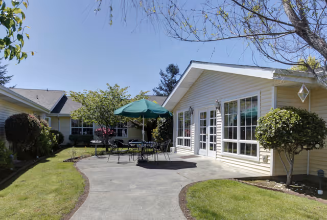 Sunny outdoor courtyard with patio tables, chairs and a green umbrella in front of a light-colored senior living building.