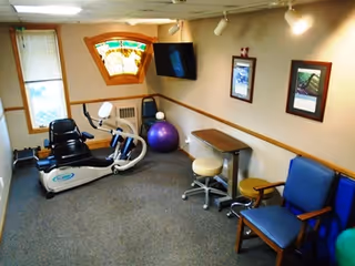 Small interior activity/therapy room with a recumbent exercise bike, exercise ball, wall-mounted TV, chairs and framed pictures.