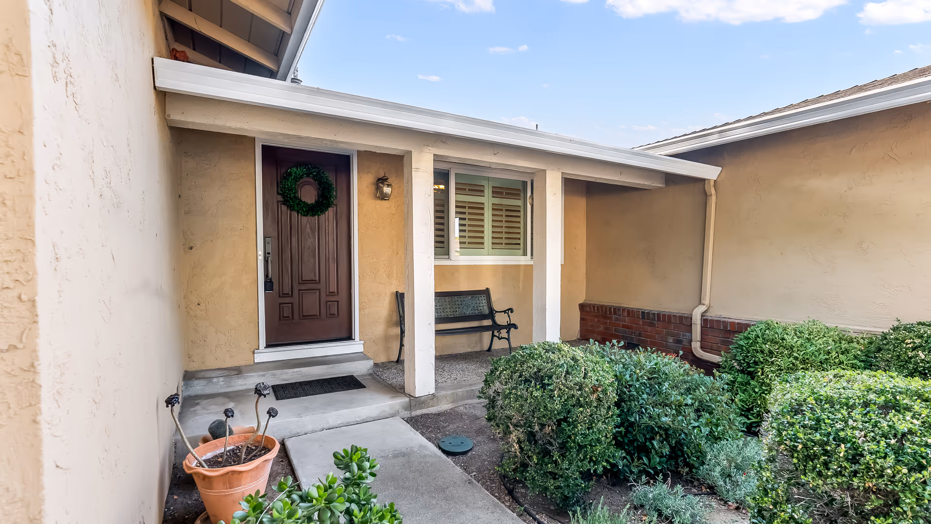 Covered entrance porch with a brown front door, a bench, and trimmed bushes along a short concrete walkway.